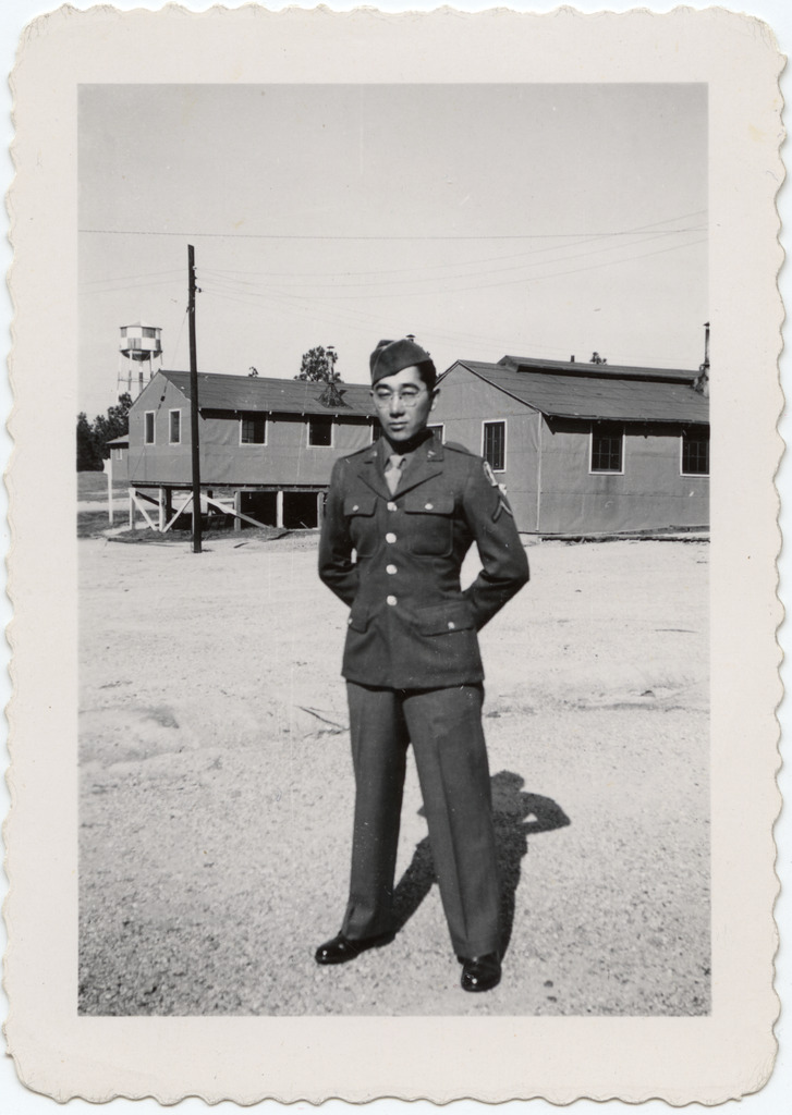 A Japanese American soldier posing for a photo in front of barracks in Camp Shelby during WWII. He is wearing an army uniform and stands with his arms behind his back.