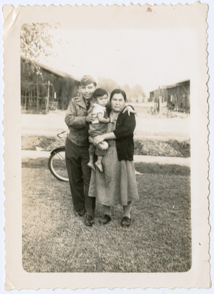 Photo of a Japanese American soldier posing with a woman and child inside Poston concentration camp. The woman is standing on the right holding up the boy, and the soldier stands on the left with his arm draped over the woman's shoulder and a hand on the boy's back. Barracks are visible in the background.