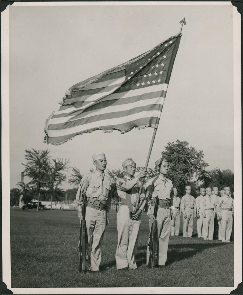 Black and white photo of three Japanese American soldiers acting as Honor Guard during a military parade. The man in the middle holds up the American flag, and two others stand on either side holding rifles against their right leg. Behind them are other soldiers standing at attention.