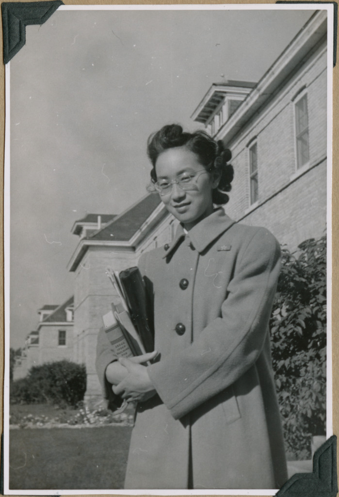 Black and white photo of a Japanese American woman standing in front of a Military Intelligence Service Language School building. She is wearing glasses and a coat, and holds several books in her arms.