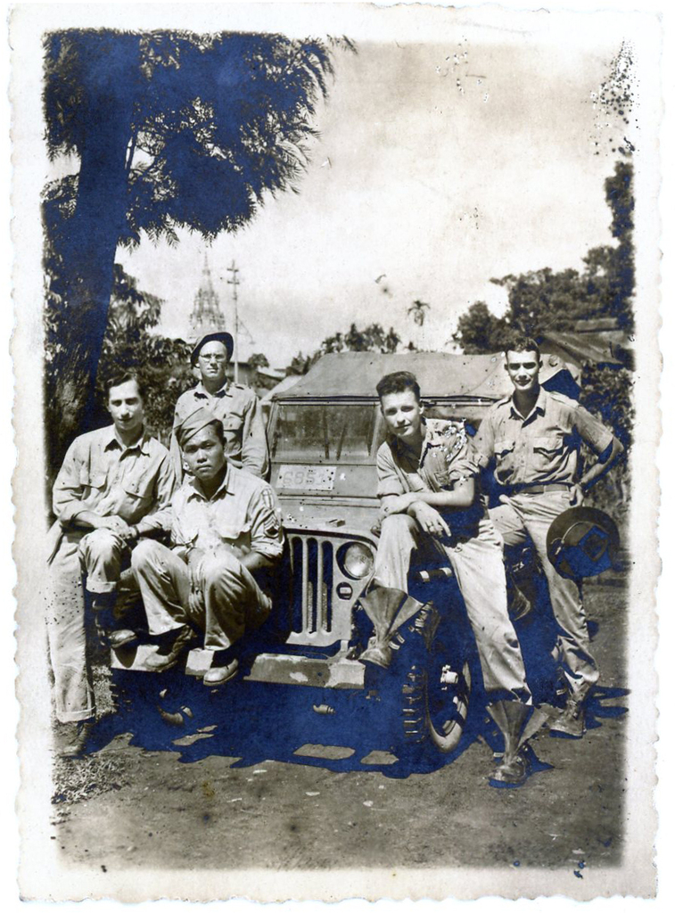 Five soldiers posing around an army Jeep. Among them is a Japanese American soldier sitting on the front of the Jeep.