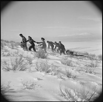Black and white photo of a group of young Japanese Americans climbing Castle Rock in the snow. Three are walking up the hill holding hands, and three more behind them are pulling a sled.
