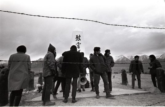 People gathered around the Manzanar cemetery monument, with barbed wire in the foreground and mountains and dramatic clouds in the distant background.