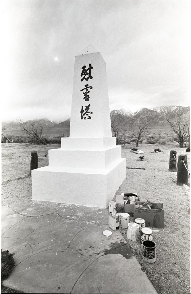 The Manzanar cemetery monument with a fresh coat of paint. It is a stone obelisk painted white with black kanji that reads "monument to console the souls of the dead." There is a box of painting supplies on the ground next to the monument, and mountains in the background.