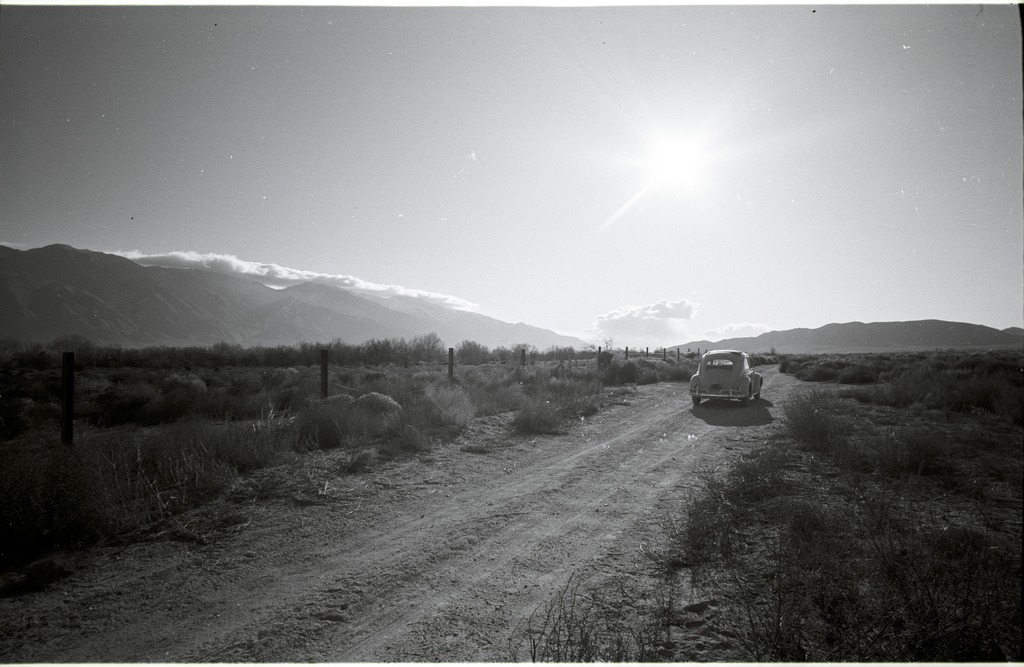 A VW Bug driving away from Manzanar after the pilgrimage. The car is on a dirt road lined with fence posts on one side.