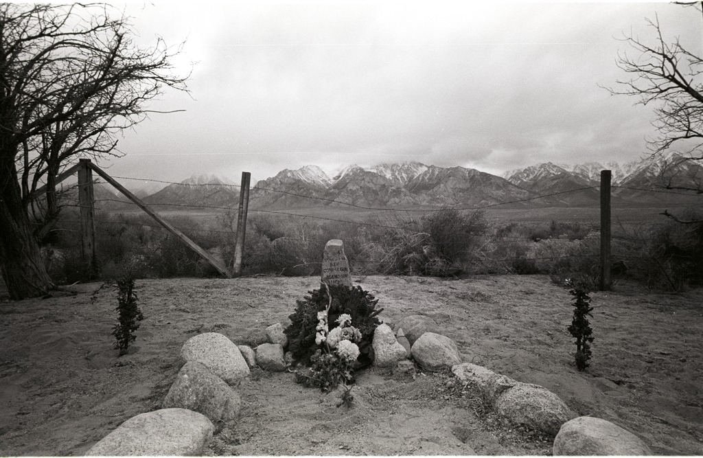 A grave site in the Manzanar cemetery. A wreath and flowers are laid in front of a grave stone, with a barbed wire fence and mountains in the background.