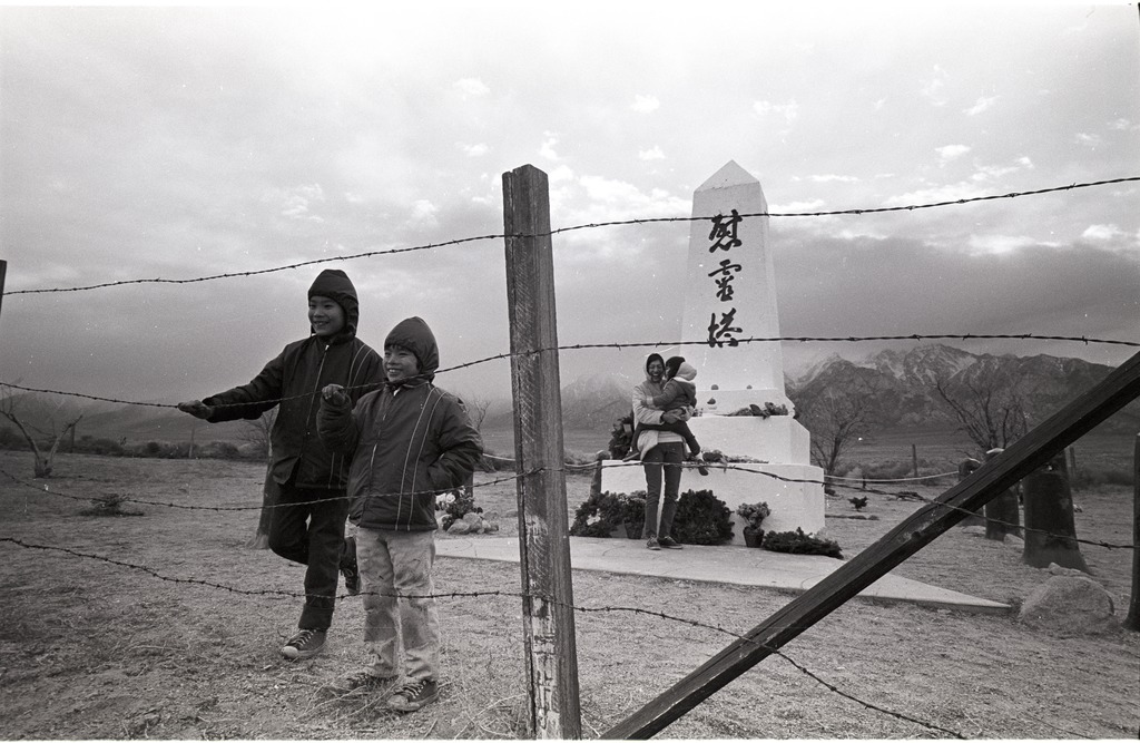 A Japanese American family at the Manzanar cemetery monument. Two preteens are touching a barbed wire fence in the foreground, and a woman is standing next to the white stone monument holding a small child behind them.