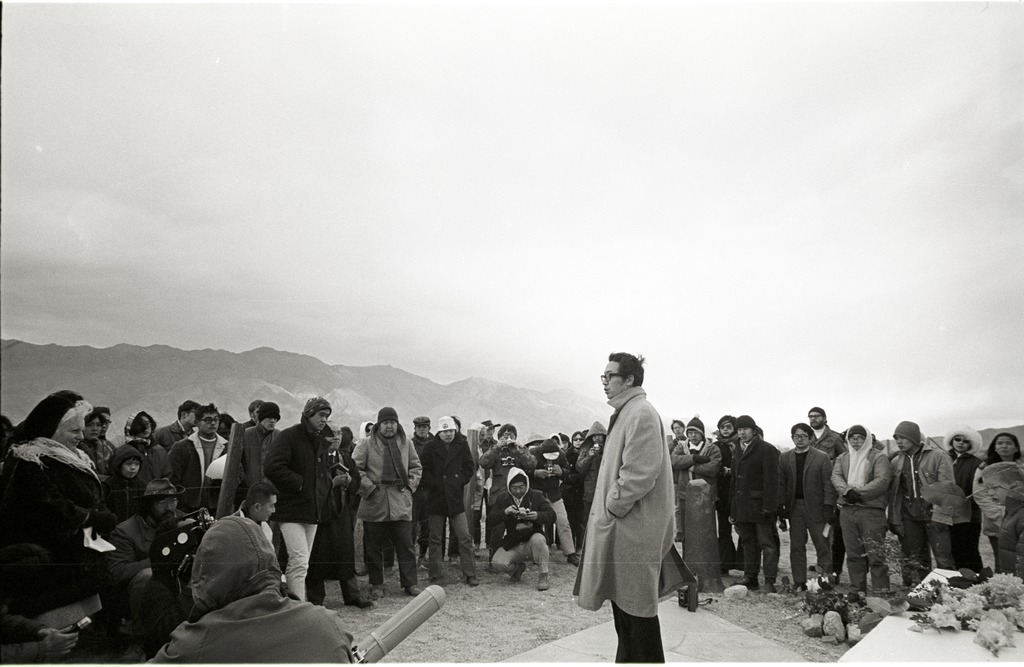 A Japanese American man wearing glasses and a heavy coat stands in the middle of a crowd of people, speaking during the pilgrimage ceremony.