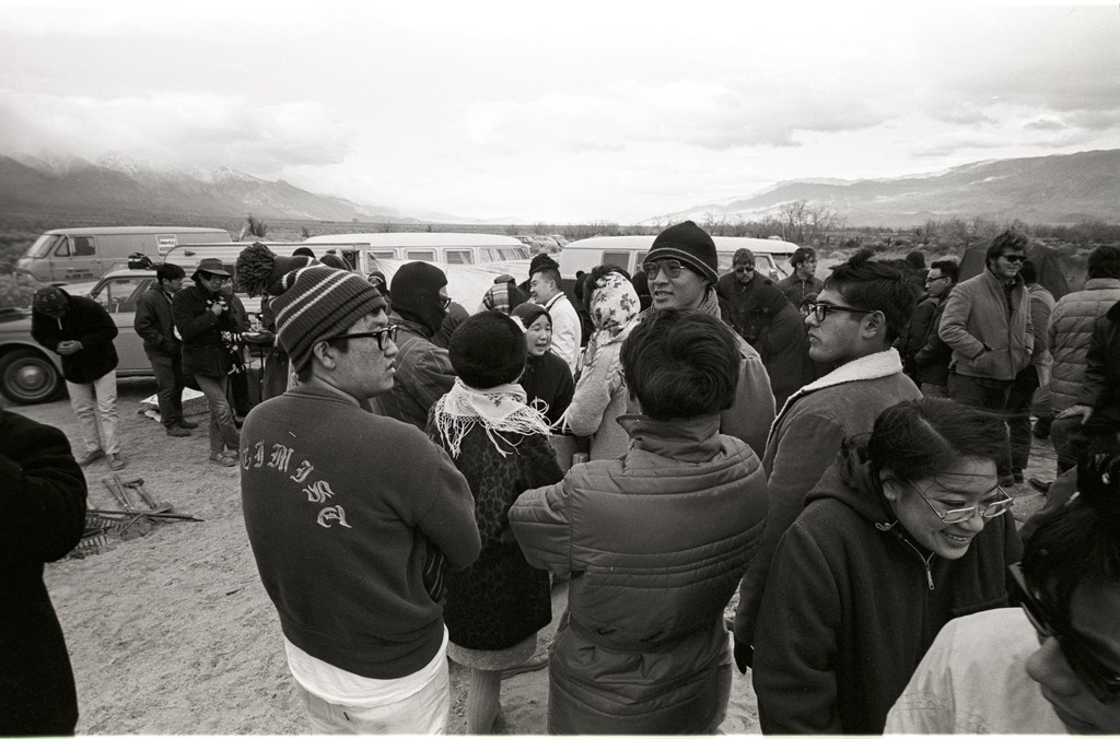 Pilgrims gathered at the former Manzanar concentration camp during the 1969 pilgrimage. There is a small group of young, college-age people in the foreground, with more people and cars in the background.