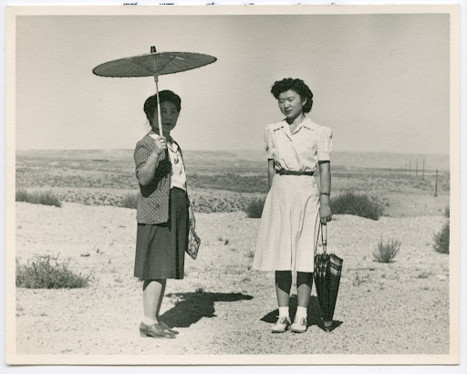 Two Japanese American women posing for a photo in the high desert near Heart Mountain. The woman at left holds a parasol over her head to shield her from the sun. The younger woman at right holds a closed umbrella at her side.