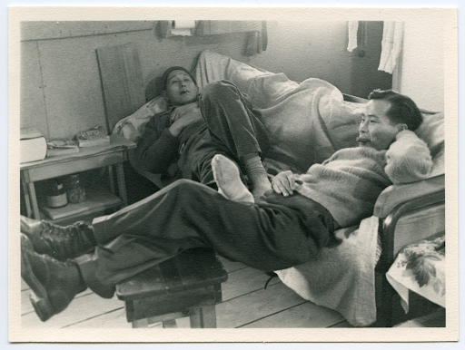 Two Japanese American men resting on a couch in a concentration camp barrack room. One man sits with his feet resting on a wooden stool, while the other is lying on the couch resting his feet on the first man's lap.
