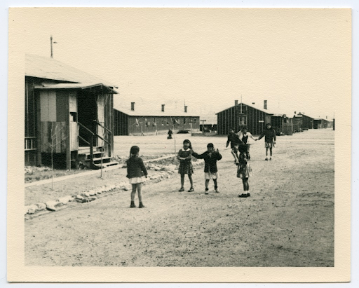 Children playing between barracks in Heart Mountain. There are four young girls in the foreground, and three slightly older girls holding hands behind them.