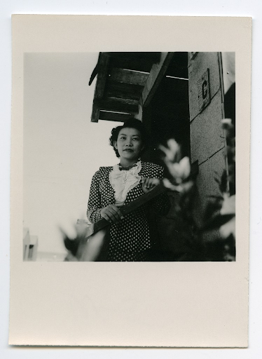 A young woman posing for a portrait on the steps in front of her barrack in Heart Mountain. Her barrack unit, C, is visible on the wall next to her.