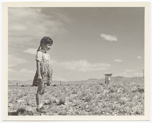 A young Japanese American girl stands in a field, with barracks and a guard tower visible in the background.