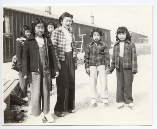 A group of Japanese American girls standing on a frozen pond in front of barracks in Heart Mountain. They are wearing ice skates and looking at the camera.