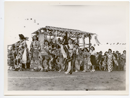 Black and white photo of women wearing kimono and dancing in front of the yagura during an Obon festival in Heart Mountain.