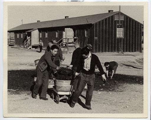 Two Japanese American men hauling coal in Heart Mountain. One is holding onto a metal tub full of coal to keep it steady, while the other pulls the wagon carrying the tub from the front. A barracks building and others gathering coal from a large pile are visible in the background.