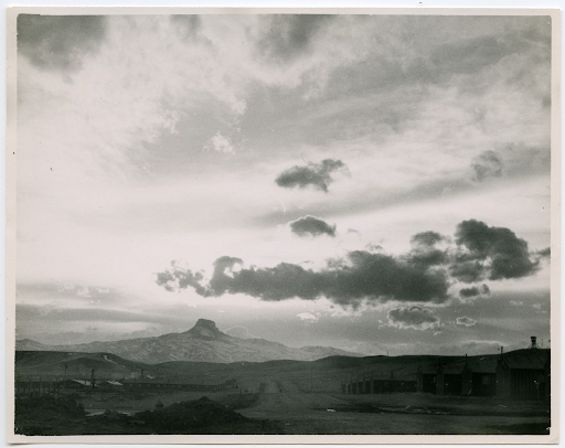 Black and white landscape photo of a sunset over Heart Mountain concentration camp. There are dramatic clouds above the mountain, which casts a shadow over barracks buildings and a guard tower.