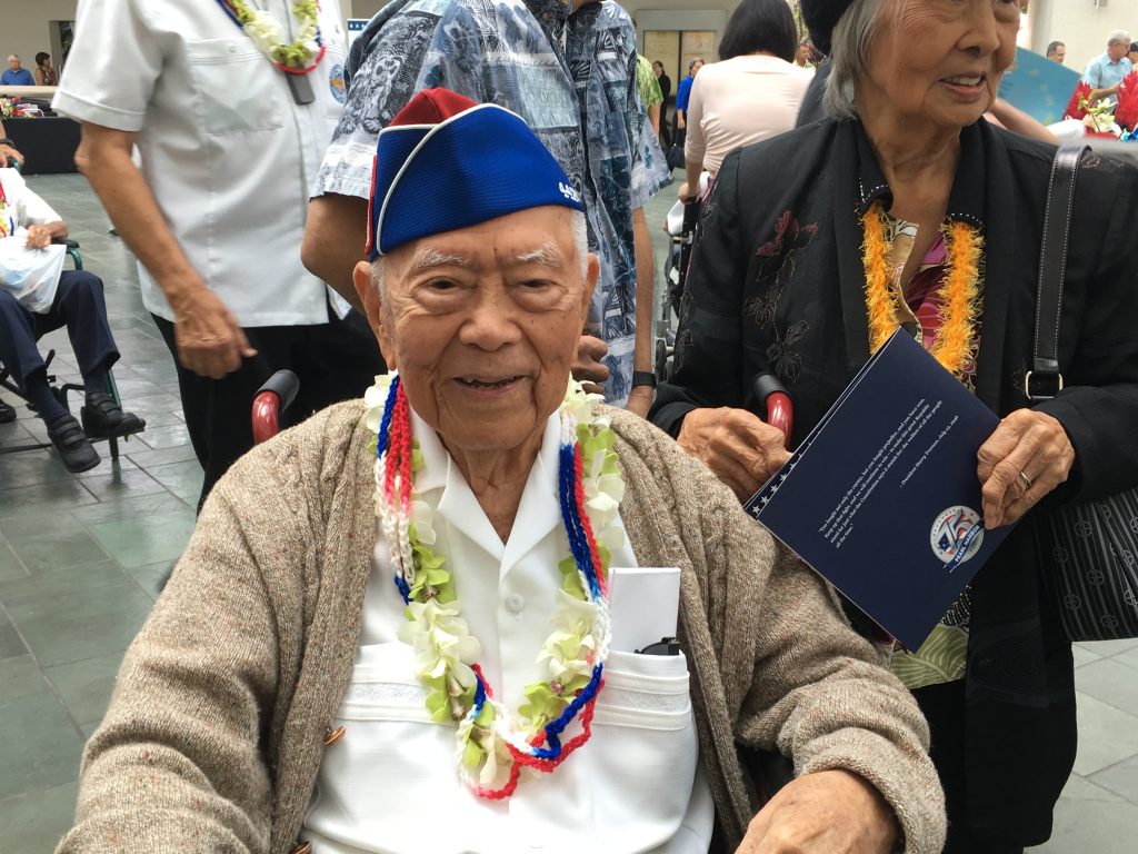 Akira Otani seated in a wheelchair at a community event. He is wearing a red and blue 442nd garrison cap, lei, and white collared shirt and beige sweater.