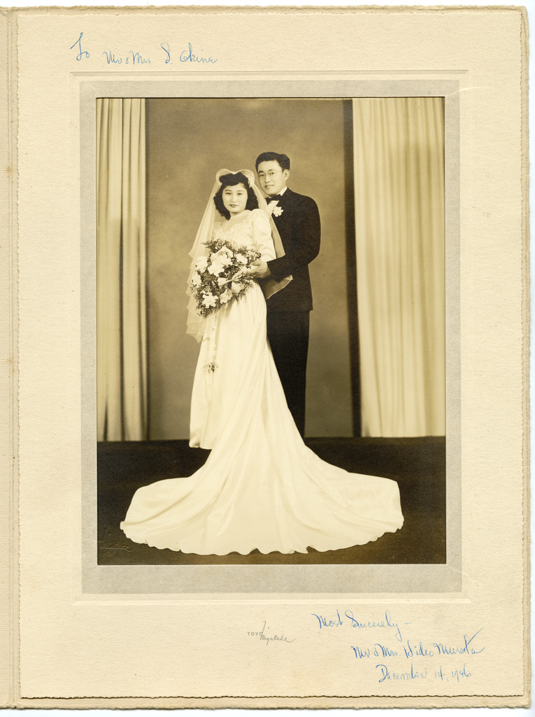 Wedding portrait of a Japanese American couple. A young woman in a veil and white silk wedding dress with a long train is holding a large bouquet of white flowers, while a man in a tuxedo with a white flower in the lapel stands behind her with his arms around her. The photo is set in a paper frame with a handwritten note that says, "To Mr. & Mrs. S. Okine, Most Sincerely - Mr. & Mrs. Hideo Murata December 14, 1946."