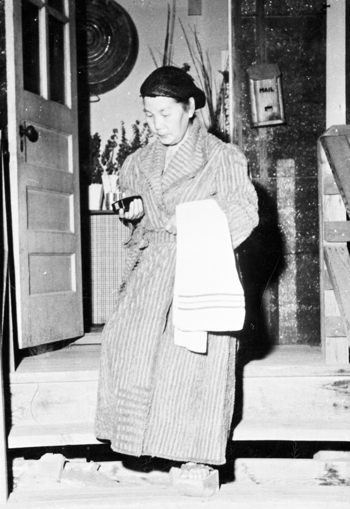 A woman stepping out of her barrack room on the way to the latrine for a shower. She is wearing a bathrobe and tall wooden geta, and carrying a towel.