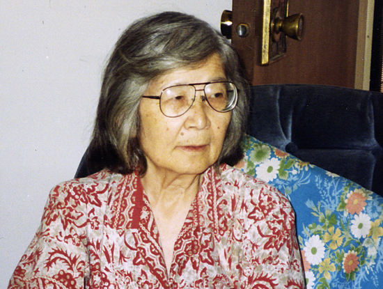 Photo of Hisaye Yamamoto from the shoulders up. She is wearing a red and white patterned blouse and sitting in a blue chair.