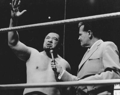 Kinji Shibuya speaking into a microphone, held by another man, in a wrestling ring. He has a "mean" look and is gesturing with one arm in the air.