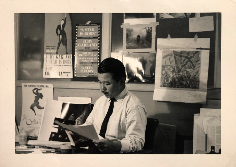 S. Neil Fujita in his studio. He is seated at a desk looking at an image on a piece of paper, next to a wall covered in posters.