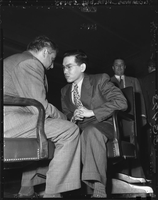 Tomoya Kawakita and his lawyer in court. The two men are sitting in leather swivel chairs facing each other and Kawakita is leaning in to listen to his lawyer.