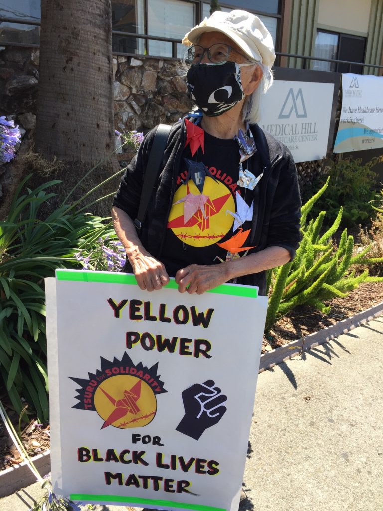 Chizu Omori holding a poster with a yellow and red Tsuru for Solidarity logo and a black fist that says "Yellow Power for Black Lives Matter." She is wearing a cloth mask and a string of folded origami cranes around her neck.