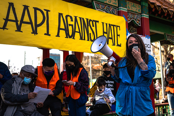 A woman holding a bullhorn and speaking at a rally. She is standing in front of a yellow banner that reads "AAPI against hate."