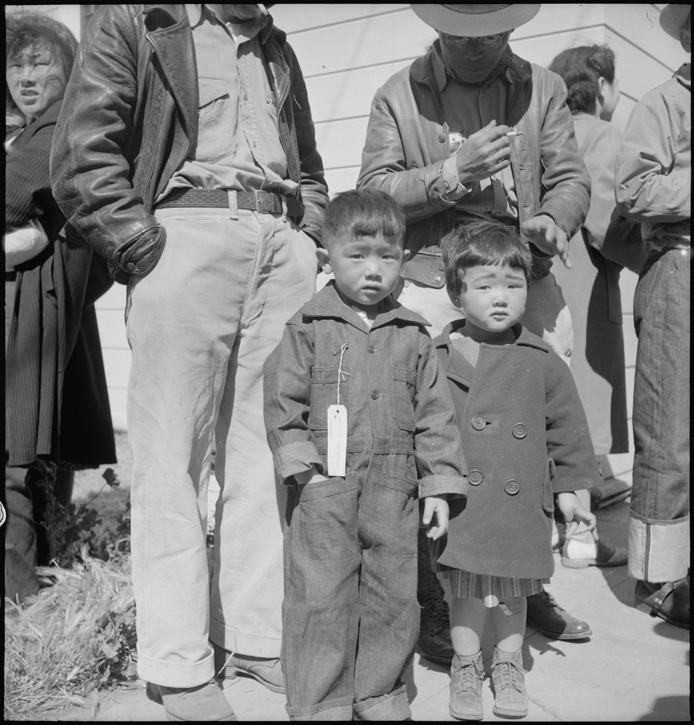 Two Japanese American children, one wearing an "evacuation" tag, standing in front of two men at a WWII incarceration camp.