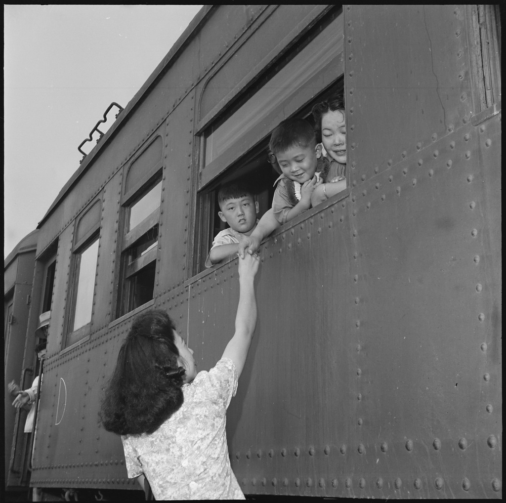 Friends saying goodbye on the train to leave Jerome. Two children are leaning out of the train window, and a woman behind them is clasping hands with a woman standing on the ground outside the train.