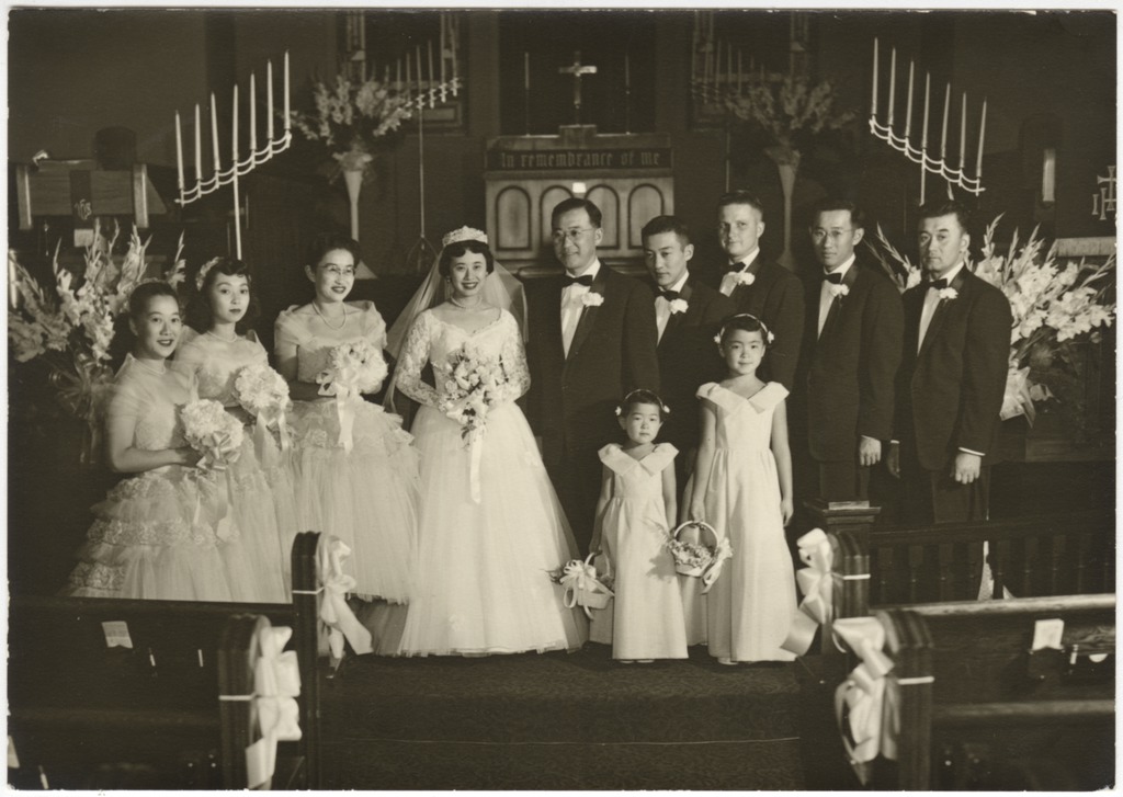 Black and white photo of a bridal party posing in front of a church altar. The bride and groom stand at the center, with three bridesmaid to the left, four groomsmen to the left, and two flower girls in front.