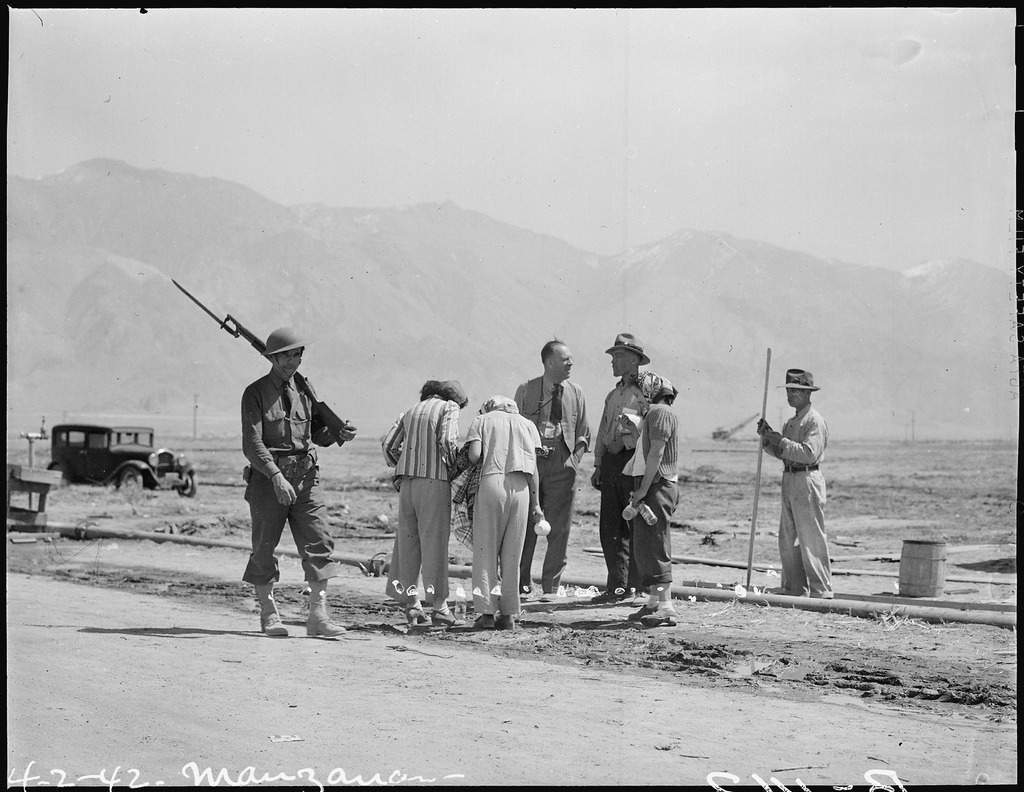 Several Japanese American men and women talking with a camp administrator at a construction site in Manzanar. A military police guard with a rifle resting on his shoulder is walking past them.