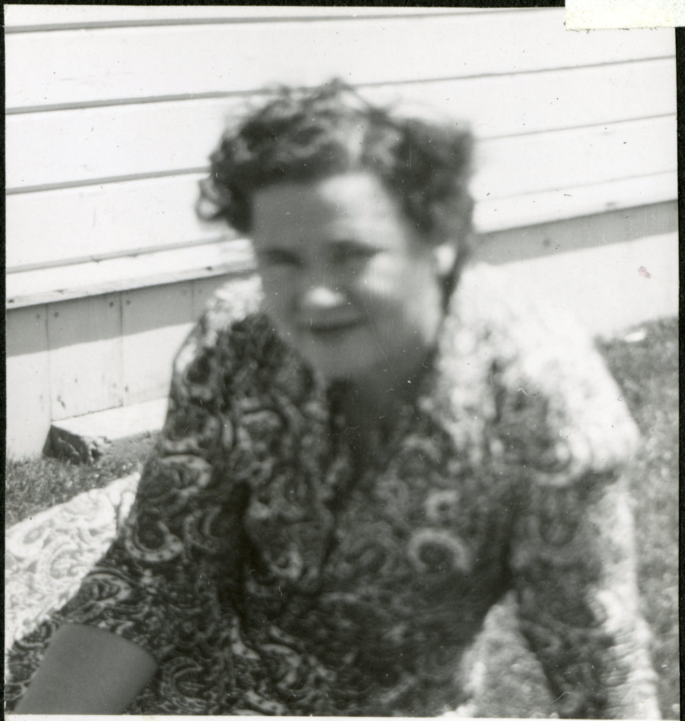 Lucy Adams sitting on a patch of grass in front of a building in Manzanar. She is wearing a paisley dress and is looking at the camera with a slight smile.