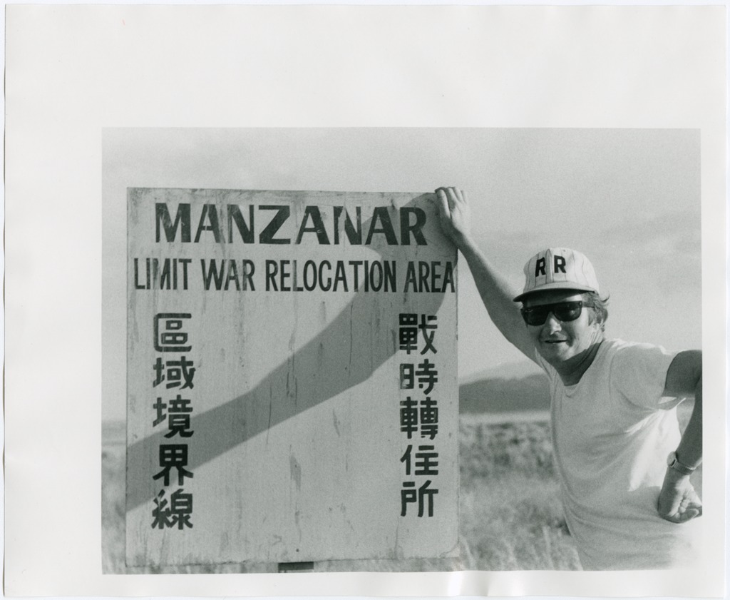 A white man wearing sunglasses, a baseball cap, and a white t shirt leans against a film prop sign that reads "Manzanar Limit War Relocation Area" in English and Japanese.