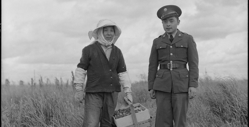 A woman farmer standing in a field next to her son. The woman is wearing protective clothing and holding a basket of strawberries, and her son is wearing a U.S. Army uniform.