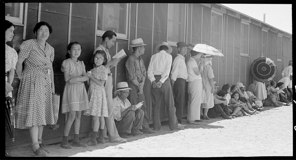 Japanese Americans waiting in a mess hall line in Manzanar. They are standing and sitting in the shade next to a barrack.