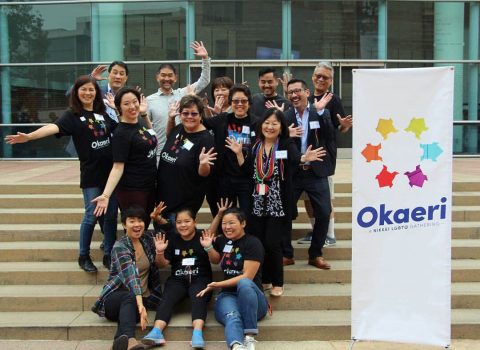 Group photo of Okaeri members smiling and waving next to an Okaeri banner