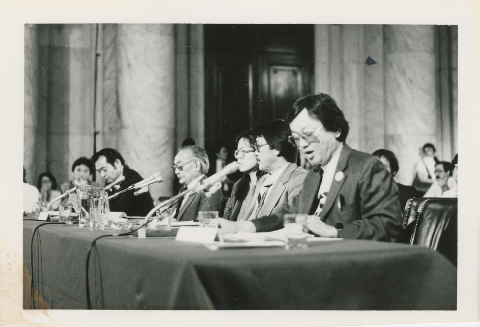 Several Japanese Americans seated at a table with microphones. Closest to the camera is William Hohri, who is reading a written statement into a microphone.