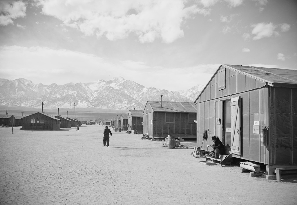 A street in between barracks at Manzanar. A man is sitting on the front steps of a barrack, and another is walking down the street. There are mountains visible in the distance.