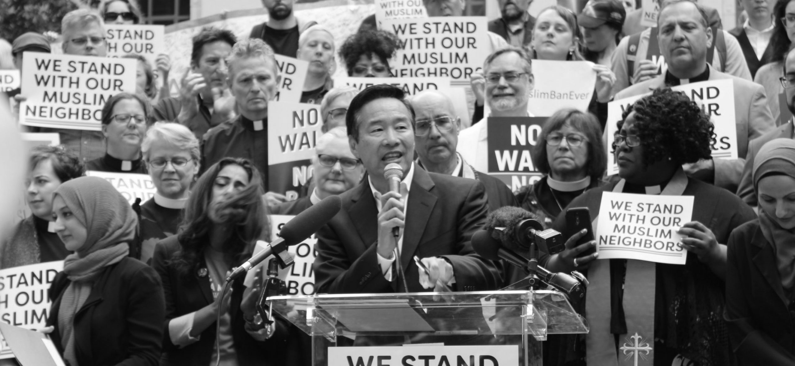 Densho Founding Executive Director Tom Ikeda speaks at a podium at a rally. Signs say "We stand with our Muslim Neighbors."