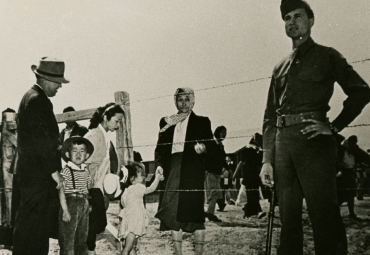 A Japanese American family stands in front of a barbed wire fence next to a guard.