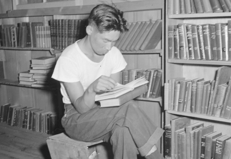 A young Japanese American man sits on a box reading in front of shelves of books.