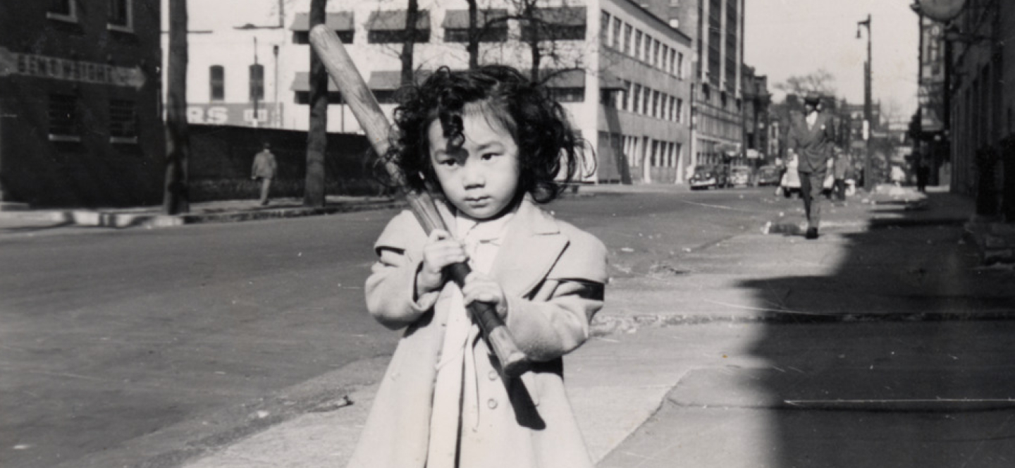 Young Japanese American girl stands on a sidewalk in a city holding a baseball bat