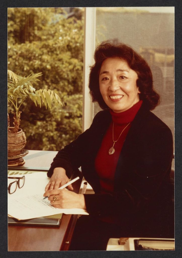 Yoshiko Uchida sitting at her desk. She is smiling and holding a pen to paper, wearing a red turtleneck sweater and gold necklace under a black blazer.