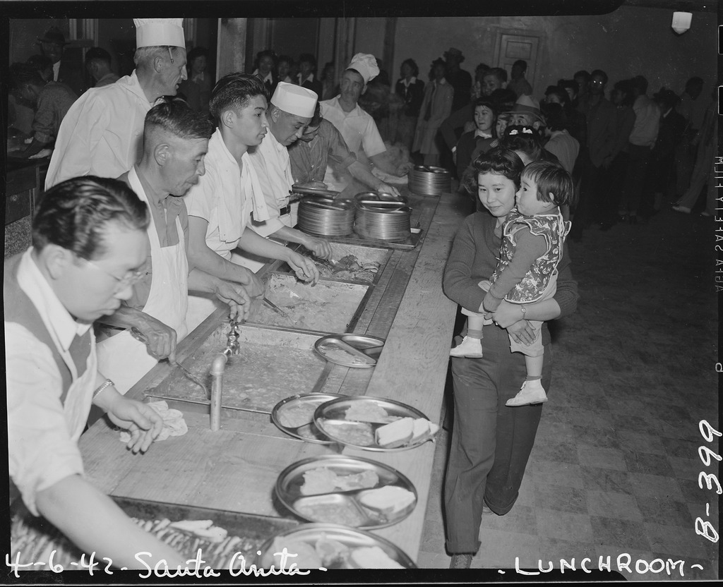 A Japanese American woman holding a small child walks past a counter where several Japanese American men are scooping food from trays onto metal plates.