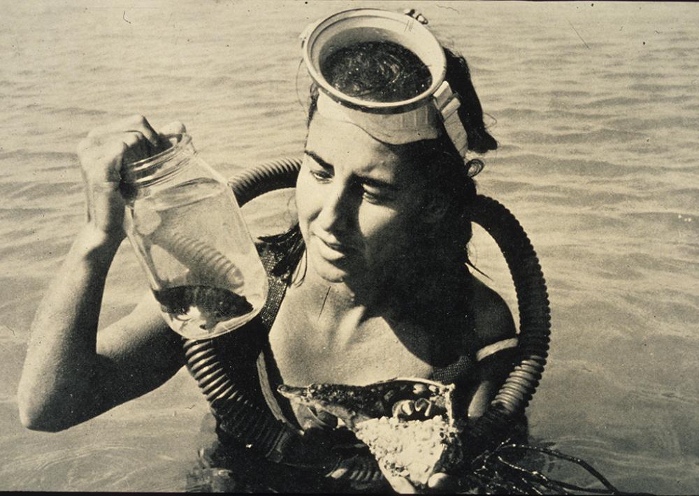 Eugenie Clark wearing scuba gear and looking at fish specimens in a glass jar.