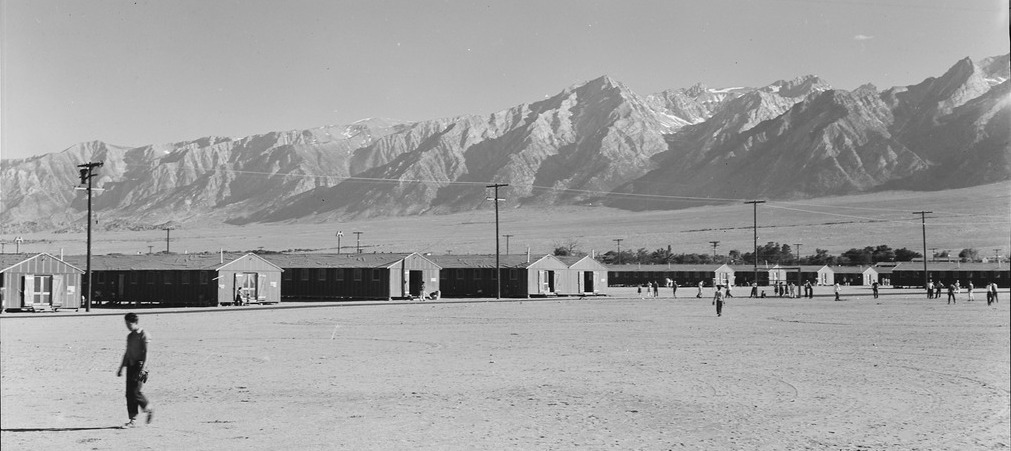 A man walking in front of barracks in Manzanar, with mountains in the distance.